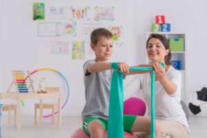 Fizjoterapeuta i chłopiec siedzący na piłce gimnastycznej ćwiczący z gumową taśmą